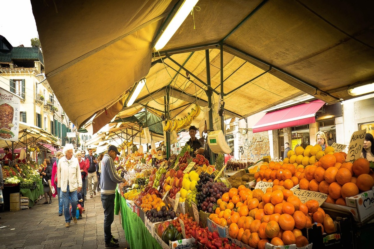 Ein belebter Markt in Sizilien mit Ständen voller frischem Gemüse wie Tomaten, Auberginen und Paprika, umgeben von einheimischen Menschen beim Einkaufen und Verkaufen.