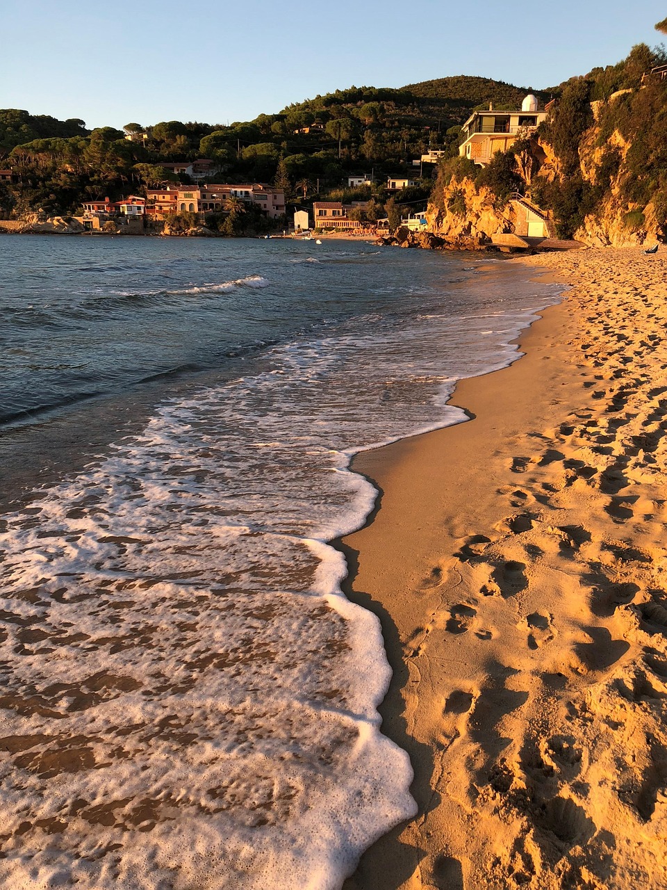 Sandstrand auf der Insel Elba bei Sonnenuntergang mit sanften Wellen, goldenem Licht, felsiger Küste und mediterranen Häusern in der Toskana.