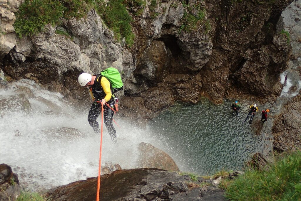 Person beim Canyoning seilt sich einen Wasserfall in einer felsigen Schlucht ab, während weitere Teilnehmer im Becken warten.