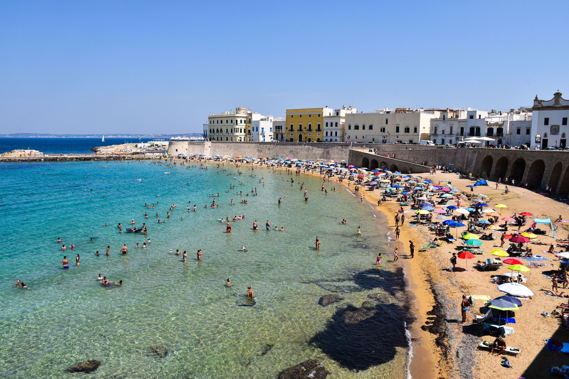 Gallipoli in Apulien mit Menschen im Wasser, blauem Meer und weißer Altstadt auf einer historischen Stadtmauer